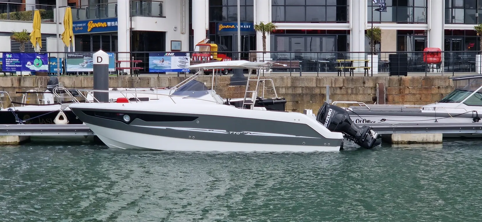 A modern boat with a grey hull docked at a marina, featuring other boats and a waterfront restaurant in the background.
